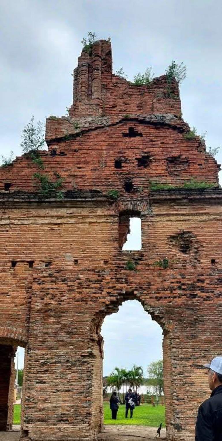Estructura de ladrillo en ruinas con vegetación, rodeada de un paisaje verde y un cuerpo de agua al fondo.