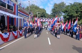 Niños marchan en formación, vestidos de uniforme oscuro, portando banderas, mientras dignatarios observan desde asientos decorativos.