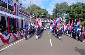 Niños marchan en formación, vestidos de uniforme oscuro, portando banderas, mientras dignatarios observan desde asientos decorativos.