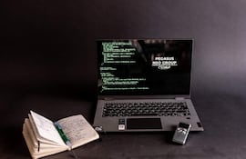 Ordenador portátil con código en pantalla oscuro, libreta y bolígrafo a un lado, sin personas presentes.
