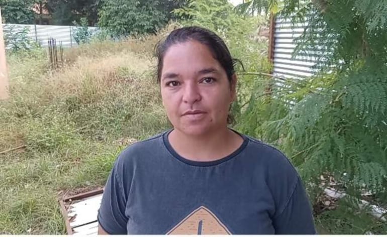 Mujer de pie en camiseta gris oscuro, con expresión comunicativa, frente a vegetación y estructura metálica.