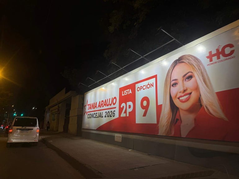 Cartel electoral de Tania Araujo con fondo rojo y sonrisa en un entorno urbano nocturno, sin personas visibles.