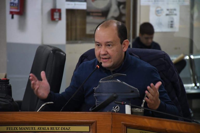 Felix Ayala en chaqueta azul, sentado en la mesa con micrófono en un evento, expresando tristeza ante la audiencia.