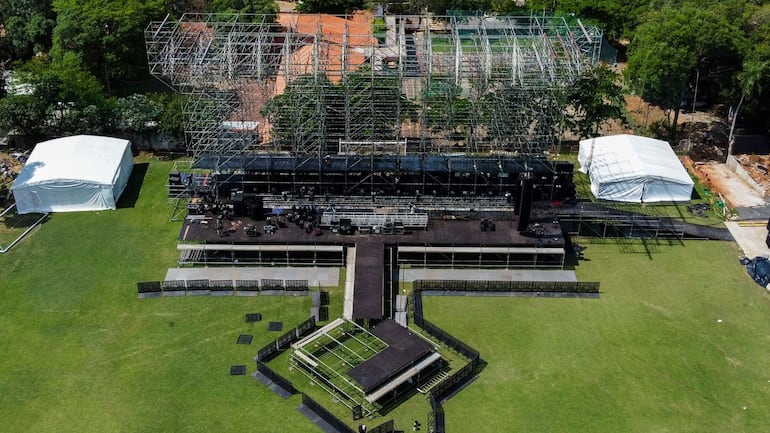 Escenario vacío con andamiaje arriba, césped verde y carpas blancas a los lados, mientras se preparan para el show.
