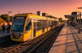 Pasajeros en estación de tren, algunos de pie y en movimiento, con un tren amarillo y el letrero 'ASUNCIÓN' visible.