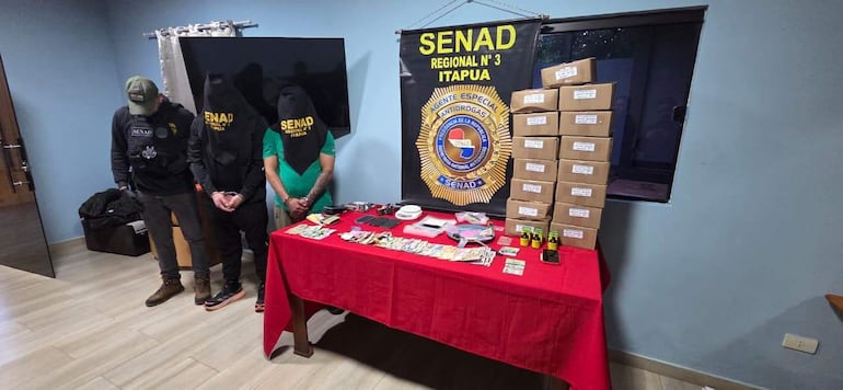 Tres hombres con capuchas en sala de la Senad, rodeados de documentos y objetos incautados en operativo antidrogas.