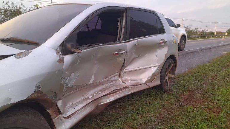 Automóvil blanco con daños en el lado izquierdo, carretera rural al fondo y otro vehículo en movimiento a la derecha.