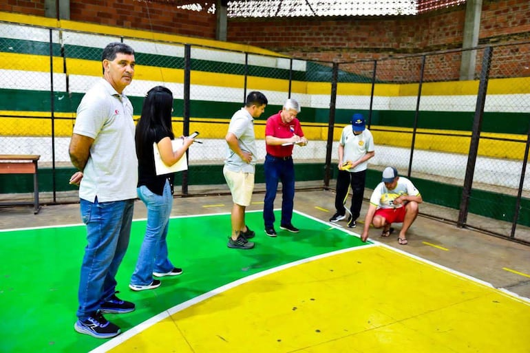 Cuatro hombres y una mujer en un recinto deportivo, con un hombre anotando y otro midiendo; destacan colores verde y amarillo en el ambiente.