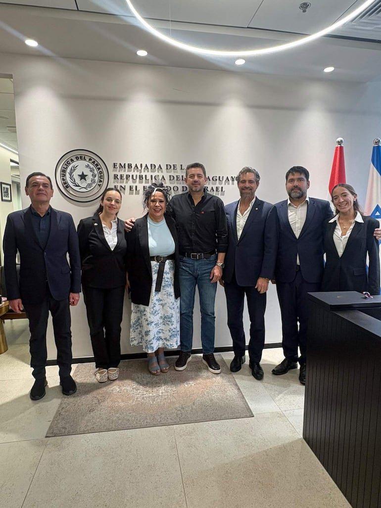Grupo de seis personas en la embajada, algunas con traje oscuro y blusas formales, sonriendo en un ambiente modernamente decorado.