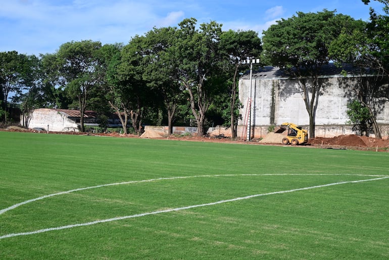 La imagen muestra un campo de fútbol en primer plano, con una portería a la izquierda y césped bien cuidado. En el fondo, hay un sitio de construcción donde se observan varias personas trabajando. Aproximadamente hay seis a ocho trabajadores, algunos de los cuales están en posiciones de pie, realizando tareas de excavación y transporte de materiales. 

No hay figuras públicas reconocibles en la imagen. En cuanto a elementos visuales que identifiquen un equipo de fútbol, no se observan camisetas, escudos o colores institucionales. 

El entorno es exterior y parece ser un área cercana a un complejo deportivo, dado que hay edificios residenciales y comerciales visibles en el fondo. Las acciones en curso incluyen la construcción y el movimiento de materiales como arena y ladrillos. No hay texto visible en la imagen.