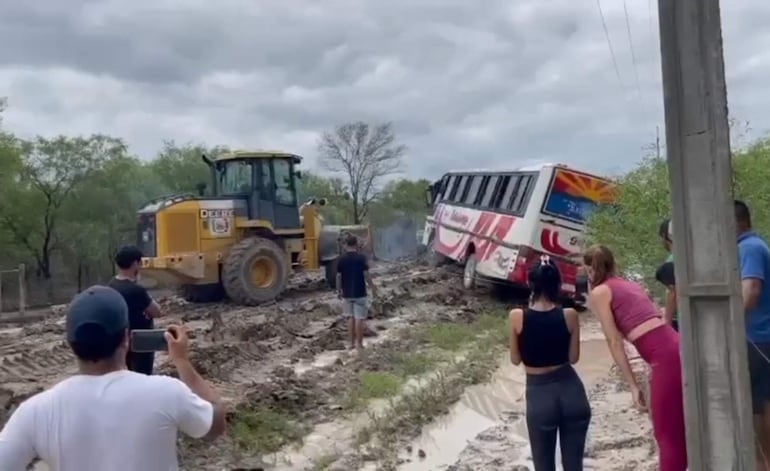 Seis personas observan un autobús atascado. Un hombre con camiseta blanca sostiene un teléfono mientras una excavadora trabaja en el fondo.