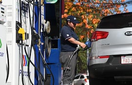 Mujer despachadora en uniforme oscuro y gorra azul, manipulando una manguera de gasolina en un día soleado.