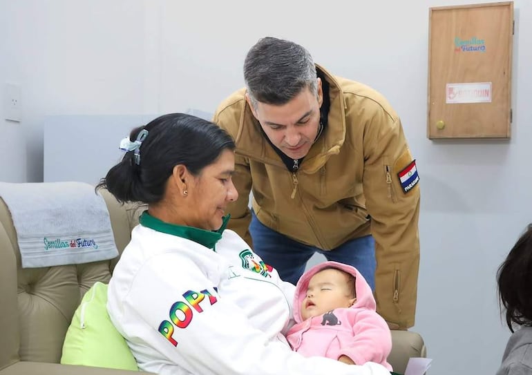 Mujer con chaqueta blanca y bebé en brazos, hombre canoso a su lado, ambiente interior con mueble y manta colorida.