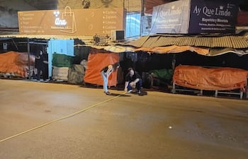 Tres personas en la calle con toldos de colores, una mide con cinta amarilla mientras observan el entorno comercial nocturno.