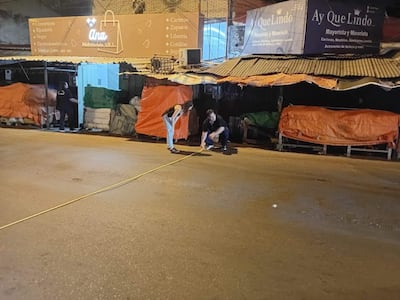 Tres personas en la calle con toldos de colores, una mide con cinta amarilla mientras observan el entorno comercial nocturno.