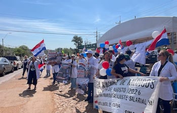 Grupo de médicos en bata blanca sostiene pancartas en manifestación en Encarnación, con banderas de Paraguay ondeando al viento.
