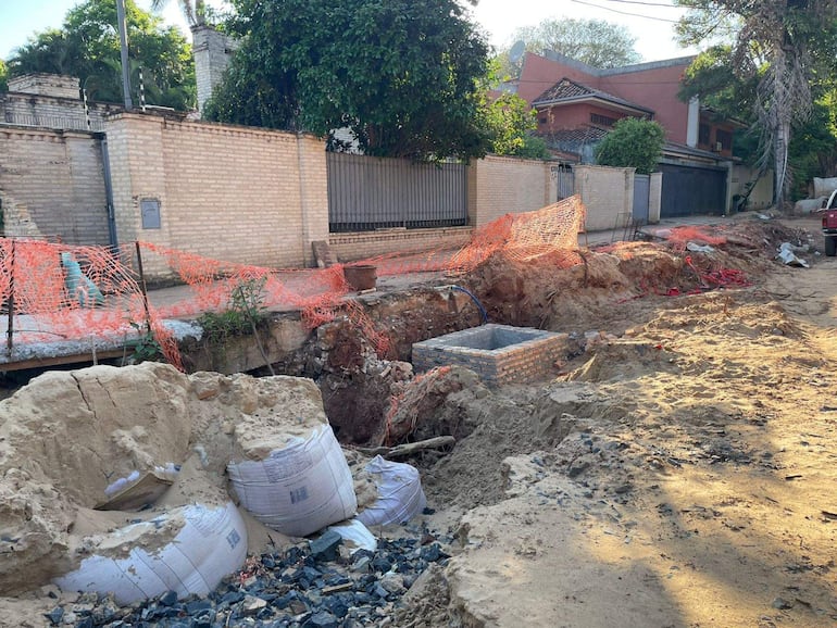 Zona de obras cerrada con malla de seguridad naranja, montículos de tierra y bolsas esparcidas en un ambiente exterior vacío.