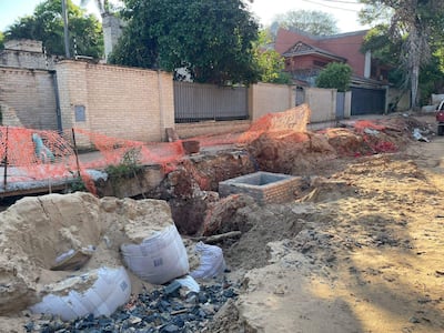 Zona de obras cerrada con malla de seguridad naranja, montículos de tierra y bolsas esparcidas en un ambiente exterior vacío.