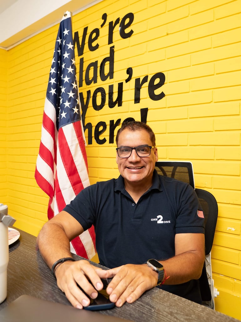 Hombre en camiseta negra con 'ONE 2 ONE', sonriendo frente a un escritorio amarillo con una bandera de Estados Unidos al lado.