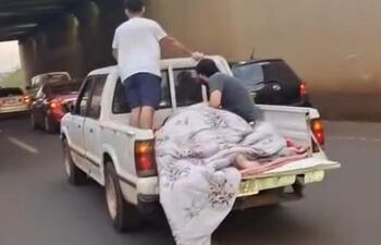 Hombre de pie y dos personas sentadas sobre manta en camioneta blanca, en un túnel con tráfico, reflejando tensión urbana.