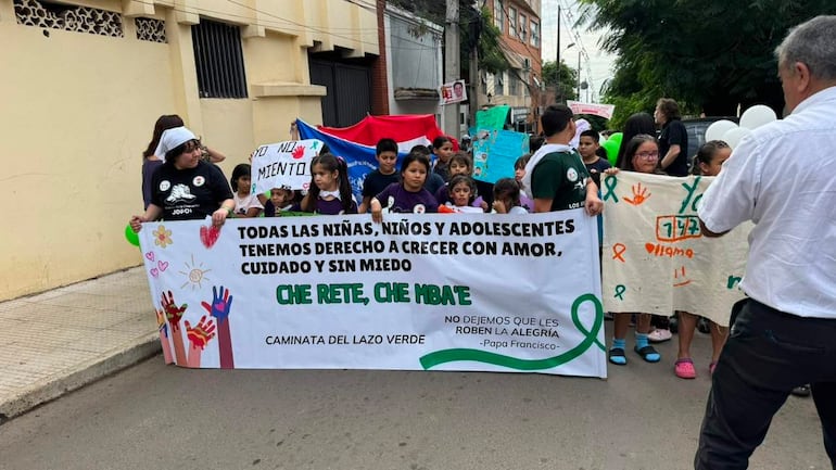 Mujer sostiene pancarta colorida en marcha, rodeada de niños con camisetas, todos en un ambiente de protesta pacífica.