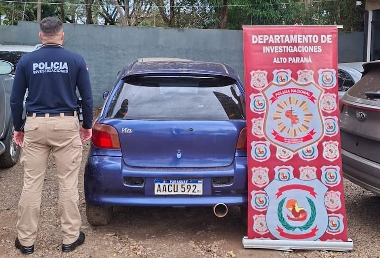 Oficial de policía de espaldas junto a un coche azul KIA Vitz, vistiendo una camiseta de 'POLICÍA INVESTIGACIONES'.