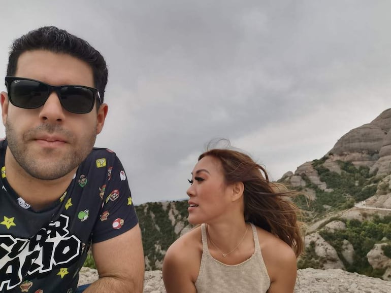 Hombre con gafas de sol y camiseta de videojuegos junto a mujer con blusa clara en un paisaje montañoso, nublado y vegetal.