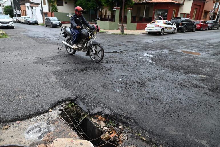 Motociclista con chaqueta negra y pantalones deportivos se aproxima a un bache en la calle, con pavimento dañado y clima nublado.