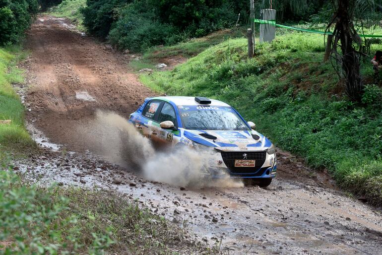 Vehículo de rally azul y blanco en movimiento, generando barro, en un camino rural con vegetación densa a los lados.