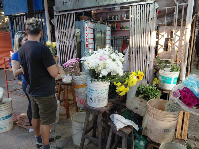 Una mujer con camiseta azul escucha a un hombre de camiseta negra que sostiene un ramo de flores amarillas en un mercado vibrante.