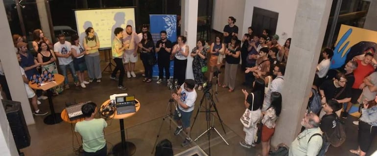Presentador vestido con camiseta colorida habla a un grupo diverso de personas en un salón iluminado, rodeado de cámaras y equipo de sonido.