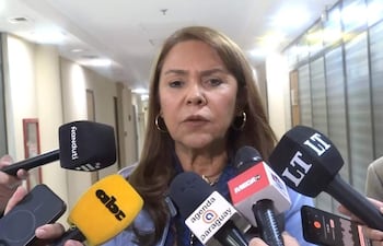 Celeste Amarilla, con chaqueta azul y aretes, habla en un podio rodeada de micrófonos de medios de comunicación.