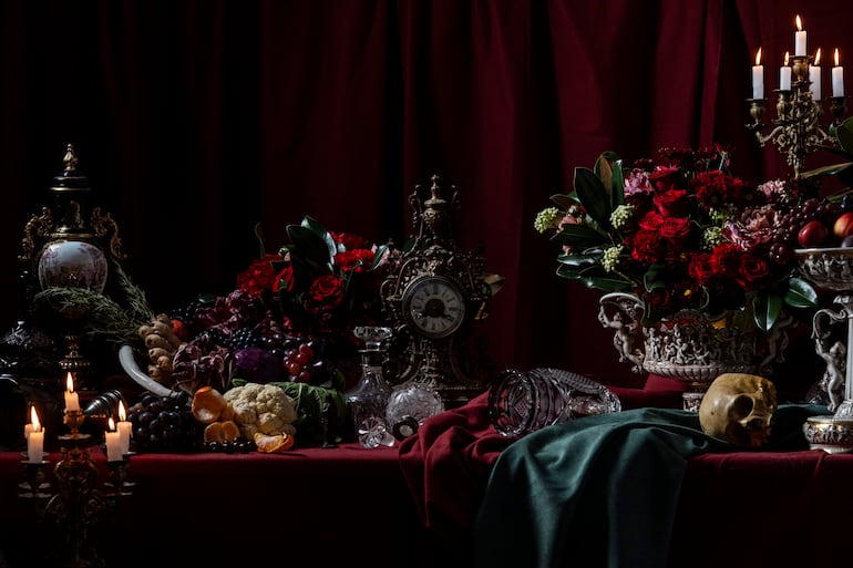 Elementos de naturaleza muerta sobre mesa roja incluyen jarrón con flores, frutas varias, reloj antiguo y candelabros con velas encendidas.