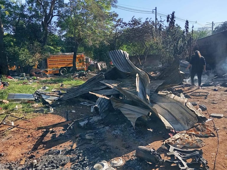 Hombre inspecciona daños en escombros de un incendio, con camión de bomberos visible detrás y humo en el ambiente.