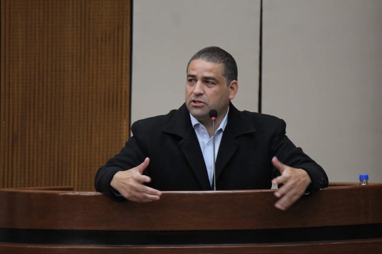 José Rodríguez en abrigo negro y camisa blanca, enfatizando su discurso en un ambiente formal con pared de madera.