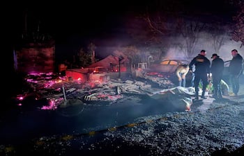 Varios hombres en ropa de emergencia trabajan en la escena de un incendio nocturno, rodeados de escombros y humo denso.