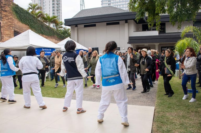 Cuatro jóvenes en trajes de taekwondo animan a un grupo de mujeres de diferentes edades en un evento al aire libre con carpas.