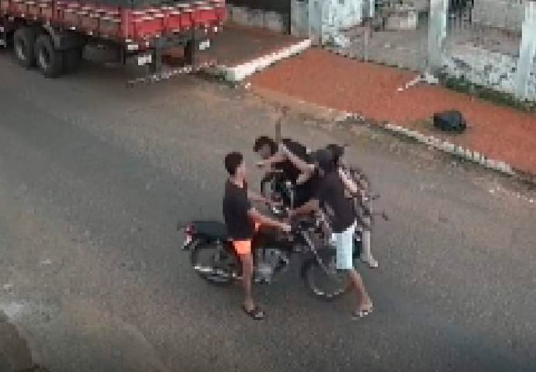 Cuatro hombres, tres en motocicletas y uno sosteniendo un objeto, visten camisetas negras y pantalones cortos de colores diversos en un entorno urbano deteriorado.