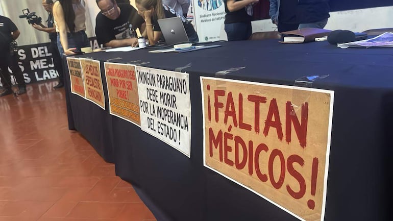 Grupo de hombres en reunión, con carteles que reclaman por falta de médicos y críticas a la inoperancia del estado.