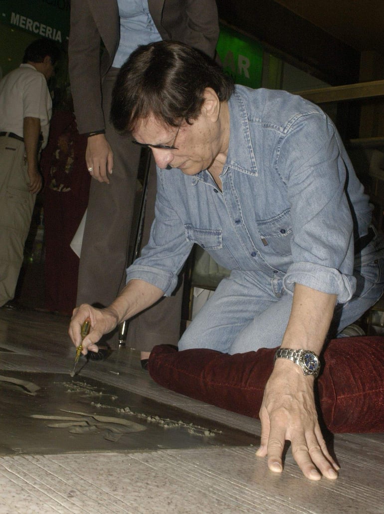 Johnny Torales agachado, con camisa de denim y anteojos, creando un relieve sobre una superficie, mientras otros observan en un entorno formal.