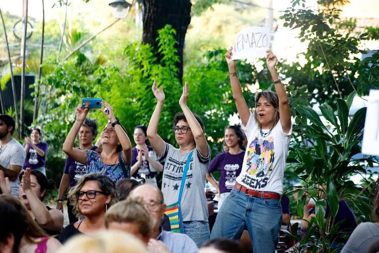 Jóvenes aplaudiendo en un parque, con una mujer en camiseta blanca y colorida levantando los brazos en celebración.