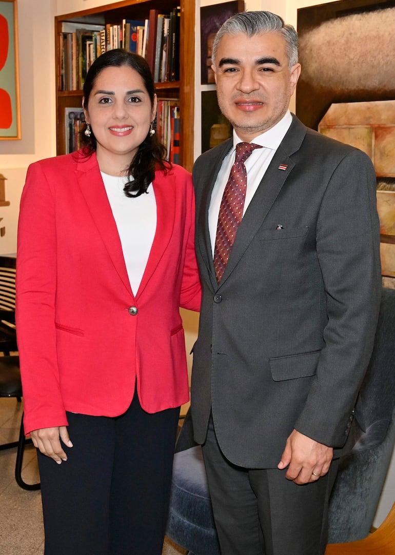 Mujer con blazer rojo y blusa blanca, sonriente, junto a un hombre en traje gris oscuro y corbata rojiza, en ambiente de oficina.