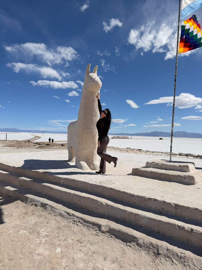 Mujer de pie con ropa oscura sonríe, tocando una escultura de llama con cielo azul y turistas de fondo.