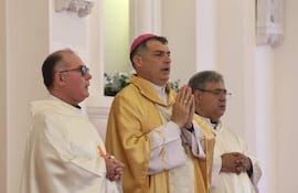 El obispo Gabriel Escobar, con mitra rosa y túnica dorada, reza junto a dos hombres con túnicas blancas en un ambiente religioso.