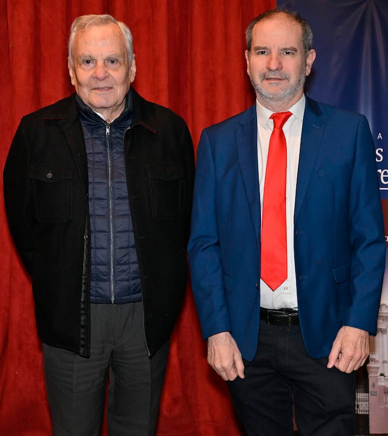 Dos hombres de pie en un evento formal; uno viste chaqueta negra y el otro traje azul con corbata roja, expresión neutra.