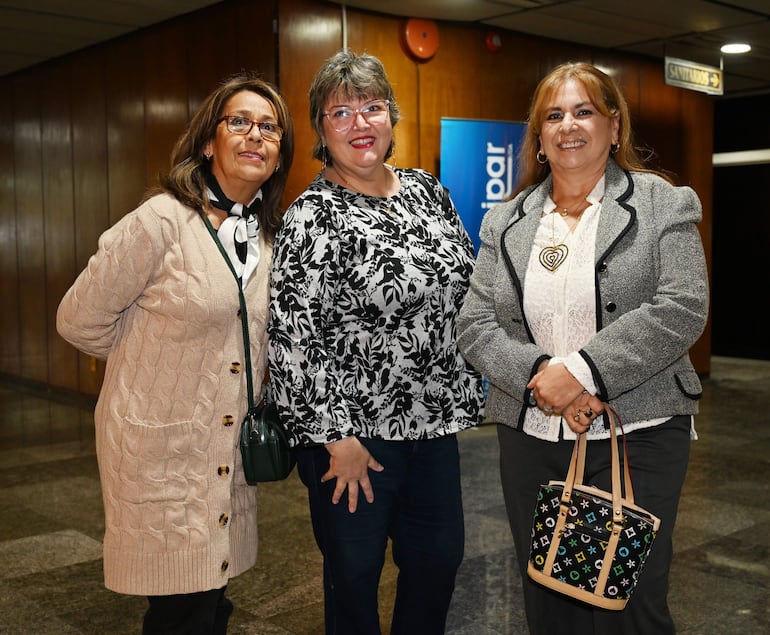Tres mujeres sonrientes: una con suéter beige, otra con blusa negra floral y la última con chaqueta gris en un ambiente acogedor.