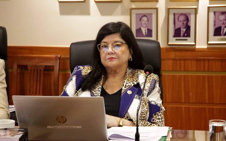 Carolina Llanes sentada en mesa de conferencias, con blusa de leopardo y chaqueta azul, prestando atención en un ambiente profesional.
