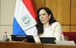Claudia Centurión, con blusa clara y abrigo neutro, habla en mesa con micrófono, una bandera de Paraguay al fondo.