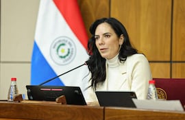 Claudia Centurión, con blusa clara y abrigo neutro, habla en mesa con micrófono, una bandera de Paraguay al fondo.
