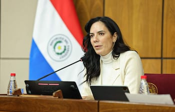 Claudia Centurión, con blusa clara y abrigo neutro, habla en mesa con micrófono, una bandera de Paraguay al fondo.